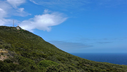 Cape Point in South Africa. A hill covered with green plants and a lighthouse at its top. Blue sky with clouds and endless ocean.