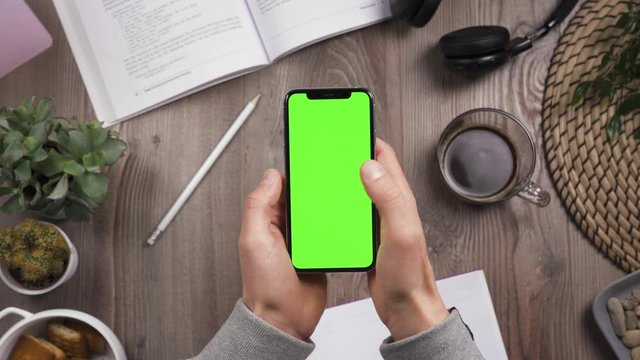 Phone with green screen chroma key in male hands. Thumb swipe left and. Top view. Workplace on wooden table with many things on the background. Daylight. POV. 4k video.