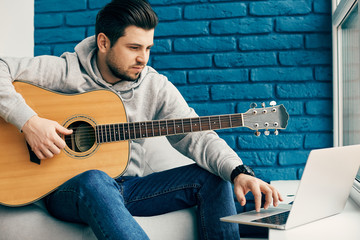 millenial with guitar sitting in modern design apartment and using laptop