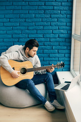 romantic man with guitar practicing at home with laptop on the bue bricks wall