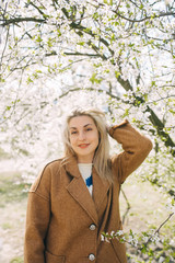 Beautiful woman in a beige coat standing in the apple garden. Vertical portrait