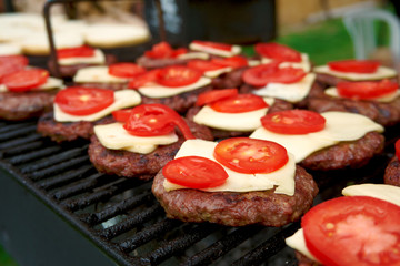 Close up of cheeseburgers prepared on grill outdoor. Garden barbecue with grilled beefburgers with cheese and tomato added. Fast food preparation in summer kitchen. Variety of food for party