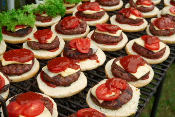 White burger buns with ketchup, beef cutlet, cheese and tomato slices placed on top. Two cheeseburgers in row with fresh lettuce leaves added. Close up of burgers placed on top of outdoor grill top