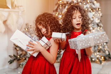 two little twin girls look at the christmas presents by the christmas tree behind