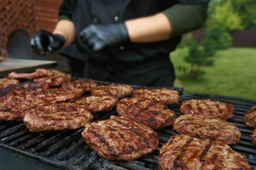 Close up of ready prepared beef burgers on grill top. Cook in background. Cook working in summer kitchen. Hamburger party outdoor during summer. Hamburger preparation during food festival. Fast food