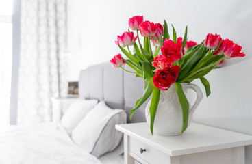 Bedroom in soft light colors..White vase with red tulips in light cozy bedroom interior. White wall, bed with white linen,