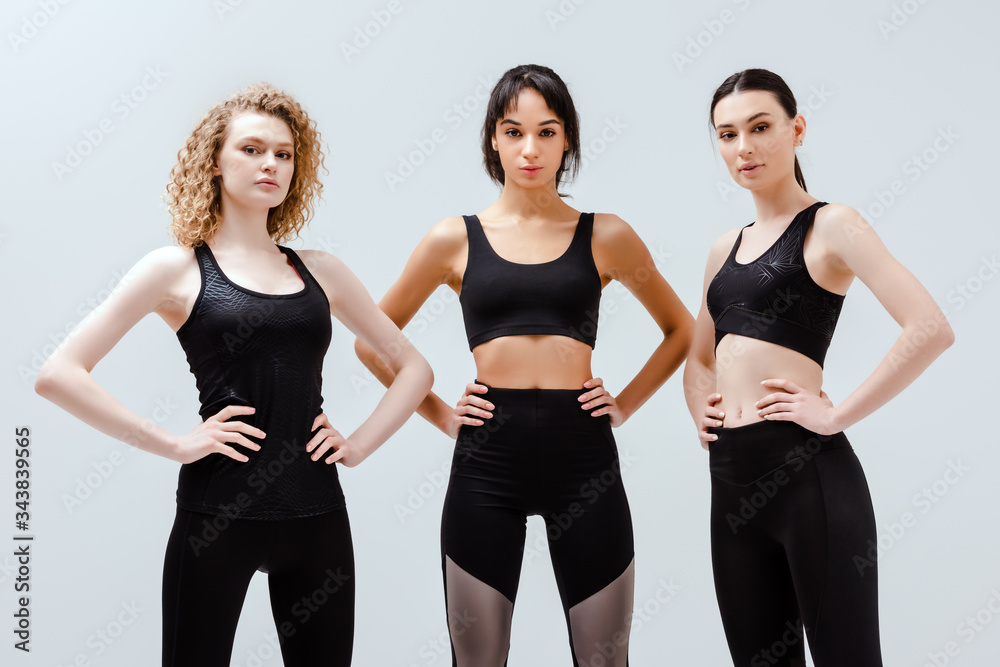 Wall mural multicultural women in sportswear standing with hands on hips isolated on white