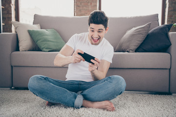 Portrait of his he nice attractive crazy addicted glad cheerful cheery guy sitting on carpet using cell playing game having fun at modern industrial loft brick interior style living-room