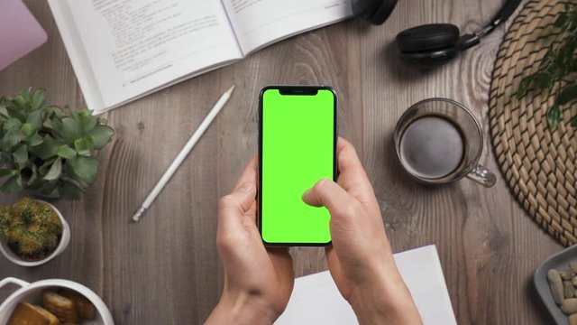 Phone with green screen chroma key in male hands. Thumb swipe up and tap likes. Top view. Workplace on wooden table with many things on the background. Daylight. POV. 4k video.