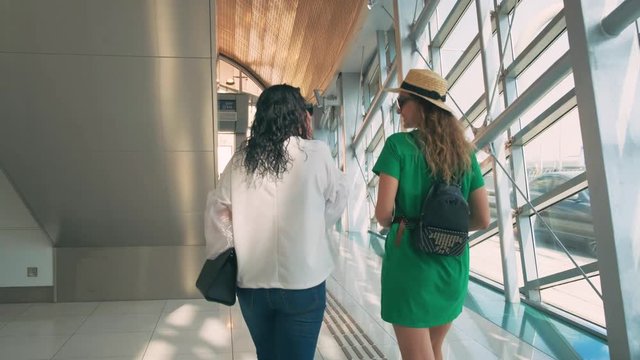 Young women tourists go pedestrian crossing. Outside window are driving cars. Girl friends turned away fun laugh. Blonde long hair. Straw hat backpack. Brunette in white blouse black bag. UAE 2020 4k