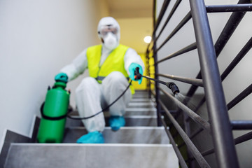 Fototapeta na wymiar Closeup of worker in sterile suit and vest sterilizing railing of a building from corona virus / covid 19. Selective focus on sprayer.