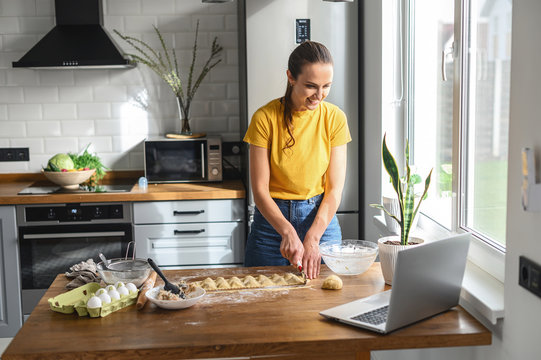 A young woman cooks at home in the kitchen, she watches a video recipe on a laptop and works with the dough on the table, she cuts ravioli. Italian cuisine at home