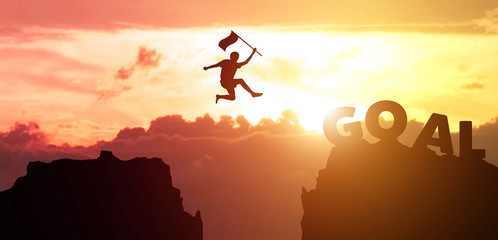 Silhouette man hand hold flag jumps from mountain to the word GOAL with sky, clouds and sunset background. Fight for reach key performance indicator in summer with copy space for label text banner.