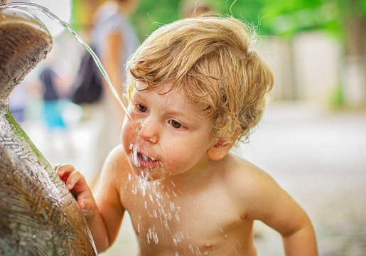 Child Drinks Water Outdoors. Little Boy Walking Around His House