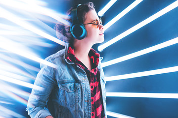 Girl stands in wireless headphones listening to music in a blue neon light. With the rays behind her.