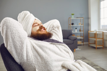 Funny bearded man in a bathrobe in a sleep mask lies in a comfortable chair in the living room. - obrazy, fototapety, plakaty