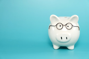 White ceramic piggy bank wearing reading glasses on blue teal background. Concept for money savings...