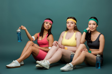 Fototapeta premium Multiethnic sportswomen sitting with sports bottles and looking at camera on green