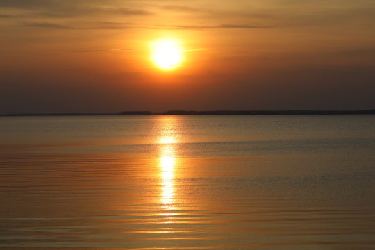 Orange sunset.The hot ball of the sun in the clear sky and the reflected Golden path on the smooth expanse of water of the lake merged on a monotonous colorful background.Calm landscape at dusk