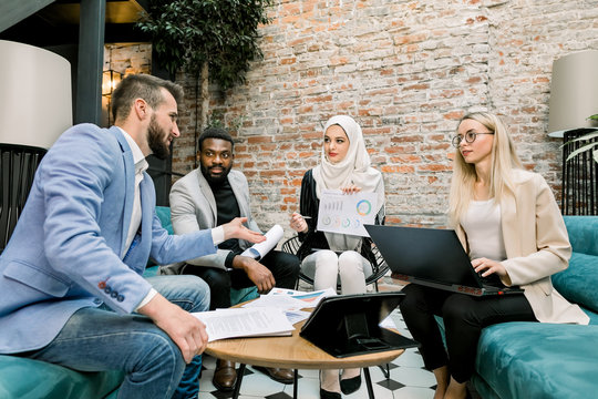 Multiethnic business people on the meeting in office. Arabian business lady in white hijab using financial document with graphs talking to her male and female partners about the results of research