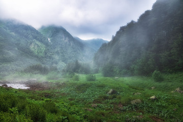 A green valley surrounded by mountains is covered in fog and clouds. Heavy fog in the mountains on a cloudy day.
