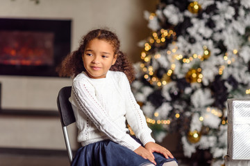 Beautiful little girl standing at the large fireplace and holding a gift, in the bright New Year's interior