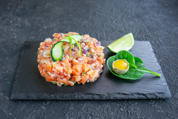 Fresh salmon tartar with quail egg, cucumber, greens and lime on black stone plate, dark table. Copy space. Seafood raw concept. Healthy, fish omega-3 rich food. 