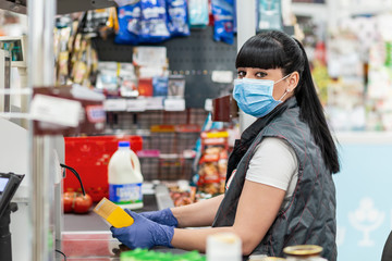 A portrait of young woman in a medical mask and gloves, working at the checkout in a supermarket....