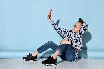 Child in sunglasses, jeans and hoodie, sneakers. Taking selfie by red smartphone, sitting sideways...