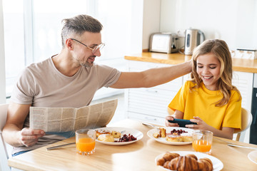 Photo of funny father and daughter using cellphone and reading newspaper