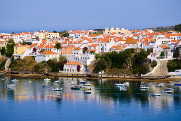Obraz premium The city Vila Nova de Milfontes is reflected in the waters of the harbor full of boats. Solo Backpacker Trekking on the Rota Vicentina and Fishermen's Trail in Alentejo, Portugal. Walking between clif