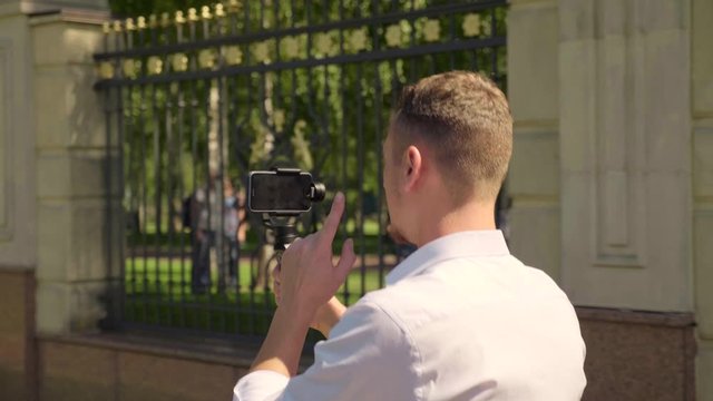Businessman-blogger is walking in the park with a stabilizer and a smartphone recording video for subscribers. A man in a white shirt is broadcasting live on a smartphone. 4K footage