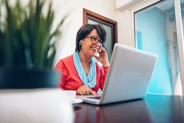 Sixty year old female teacher wearing headphones having online class via video chat on laptop computer. She is sitting on a wooden modern desk at home. Smiling and enjoying communication