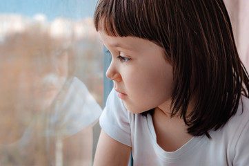 a little girl child at home during quarantine looks hopefully through the window glass.