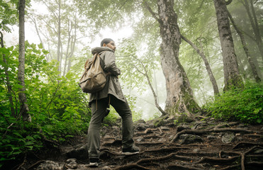 A man in a foggy forest with a backpack and in a rain jacket turned around. Travel and the environment. Human and nature.