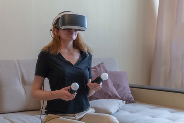 Beautiful young woman with virtual reality headset and joysticks playing video games home sitting on the sofa.