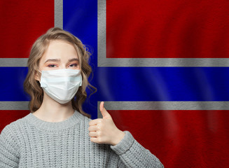 Happy woman in face mask holding thumb up on Norway  flag background. Flu epidemic and virus protection concept