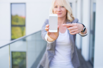 Mockup blonde woman holding her smartphone with empty screen in a modern building. Real Estate Agent with mockup smartphone. Young woman with phone in modern background