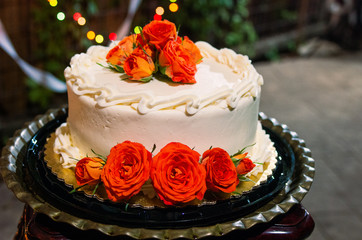 White creamed icing cake decorated with orange flowers and colored lights in background.