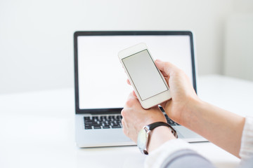 Close-up of smartphone with blank screen in hands of young woman sitting at white table and touching screen. On table laptop with blank screen. White style. Minimalism. Mock up,space for ad