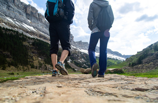 View from behind of a couple walking on a dirt road between snowy rocky mountains with a cloudy sky.