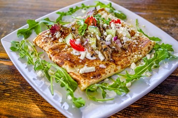 Greek salad crepe sandwich with arugula, tomatoes, cucumber, and feta cheese on white decorated plate.