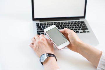 Close-up of smartphone with blank screen in hands of young woman sitting at white table and touching screen. On table laptop with blank screen. White style. Minimalism. Mock up,space for ad