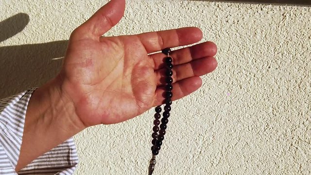 Slow motion. Dark-skinned male hands holding aosary folded in prayer gesture. Close-up. Muslim man in light shirt makes prayer. Sunlight, sunny day. - Powered by Adobe