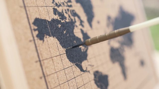A red and blue darts dart arrow with his point sticking in a cork board world map  where he landed in the middle of the United states of America and Canada on the American Continent. Extreme closeup