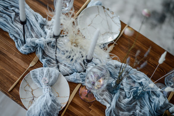 Minimalistic table setting for holiday dinner, wooden table with dried flower, plates, golden cutlery, white candeles, bright blue table runner. Selective focus. Top view