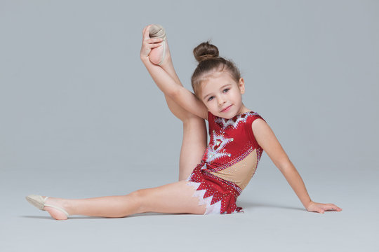 Flexible little girl in beautiful red dress is stretching doing gymnastic exercises on grey background
