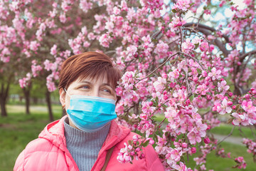 portrait of adult sad woman of risk group in medical mask outdoor by the blooming pink apple tree in spring. Quarantine, coronavirus, spring, age concept