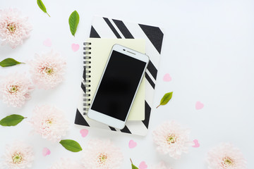 white smartphone with a black screen on yellow and black and white notepads and many pink flowers of chrysanthemums and green leaves on a white table. space for text...