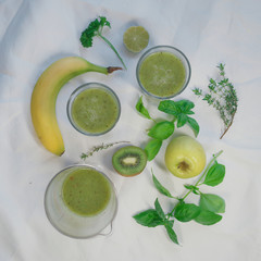 Still life in green tones on a table with a white tablecloth with glasses with green juice of parsley and ginger kiwi and fruits, banana and green apple